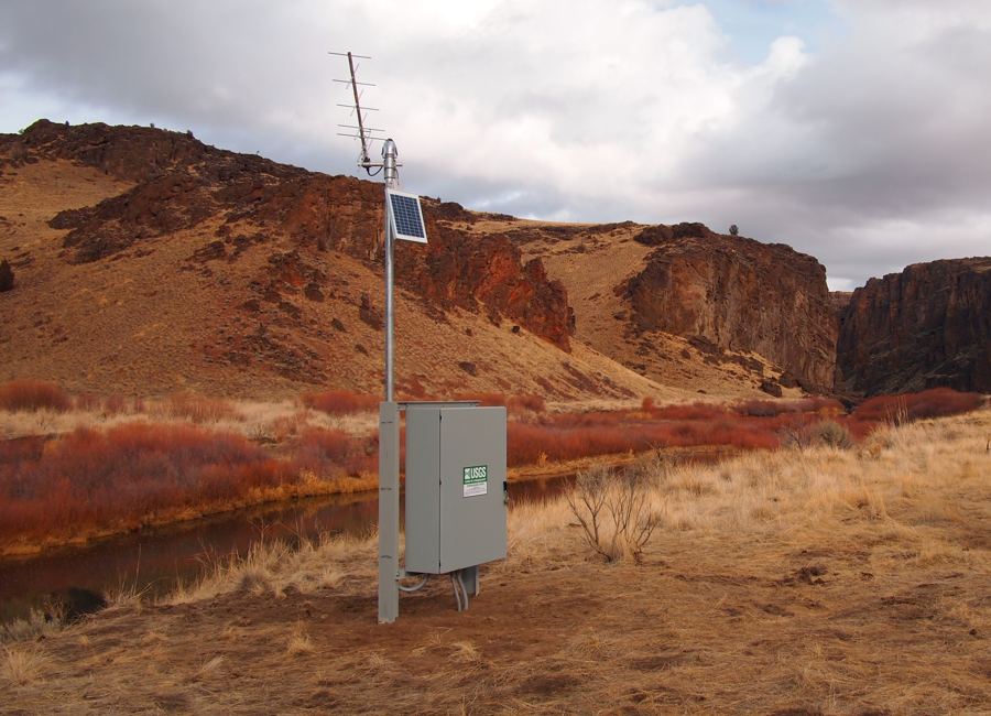 A USGS streamgage with an antenna, solar panel, and equipment cabinet stands beside the East Fork Owyhee River below red canyon walls.